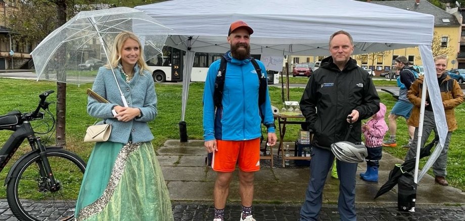 Bei etwas regnerischem Wetter stehen Loreley-Repräsentantin Katharina Blanckard, Läufer Simon Fischer und Bürgermeister Mike Weiland vor einem Pavillon in den Rheinanlagen in St. Goarshausen. 