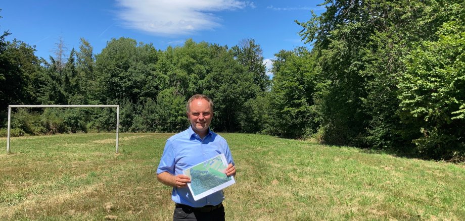 Bürgermeister Mike Weiland steht bei sommerlichen Wetter auf einer grünen Wiese, die als zukünftiger Standort des Mobilfunkmastes in Hinterwald vorgesehen ist. Im Hintergrund sind ein Fußballtor und Bäume zu sehen.