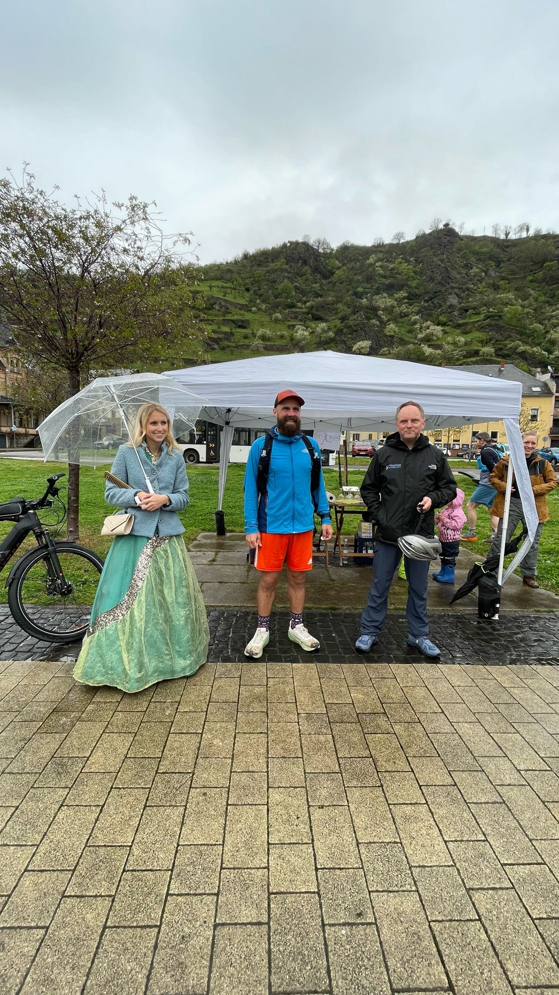 Bei etwas regnerischem Wetter stehen Loreley-Repräsentantin Katharina Blanckard, Läufer Simon Fischer und Bürgermeister Mike Weiland vor einem Pavillon in den Rheinanlagen in St. Goarshausen. 