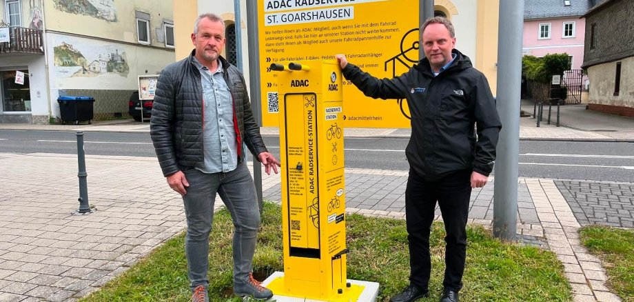 Foto Radreparatursäule Verwaltungsmitarbeiter Armin Schaust und Bürgermeister Mike Weiland stehen neben der neben der Radreparatursäule. Dahinter befindet sich ein ebenfalls gelbes Schild, dass auf die Zusammenarbeit mit dem ADAC-Mittelrhein hinweist. Im Hintergrund sind die Straße und weitere Gebäude zu sehen.