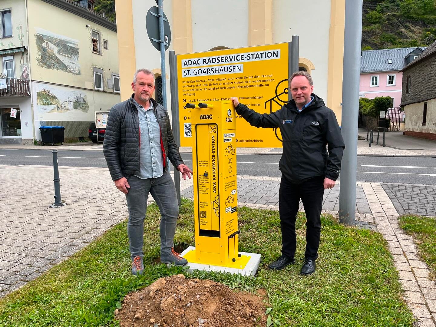 Verwaltungsmitarbeiter Armin Schaust und Bürgermeister Mike Weiland stehen neben der neben der Radreparatursäule.  Dahinter befindet sich ein ebenfalls gelbes Schild, dass auf die Zusammenarbeit mit dem ADAC-Mittelrhein hinweist. Im Hintergrund sind die Straße und weitere Gebäude zu sehen.