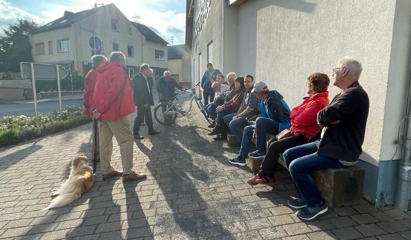 Etwa 15 Menschen sitzen nebeneinander auf einer Bank. Ein paar weitere stehen neben Bürgermeister Mike Weiland, der gerade etwas am vortragen ist.
