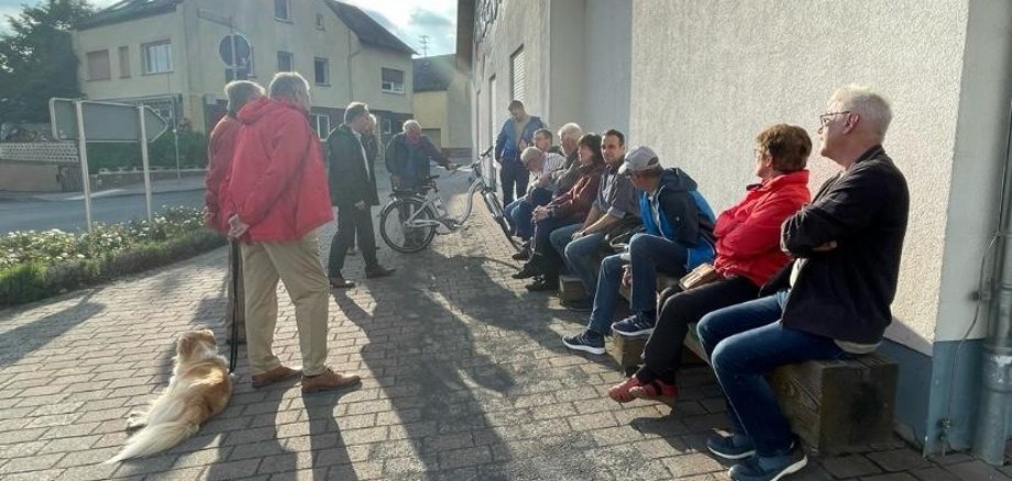 Etwa 15 Menschen sitzen nebeneinander auf einer Bank. Ein paar weitere stehen neben Bürgermeister Mike Weiland, der gerade etwas am vortragen ist.