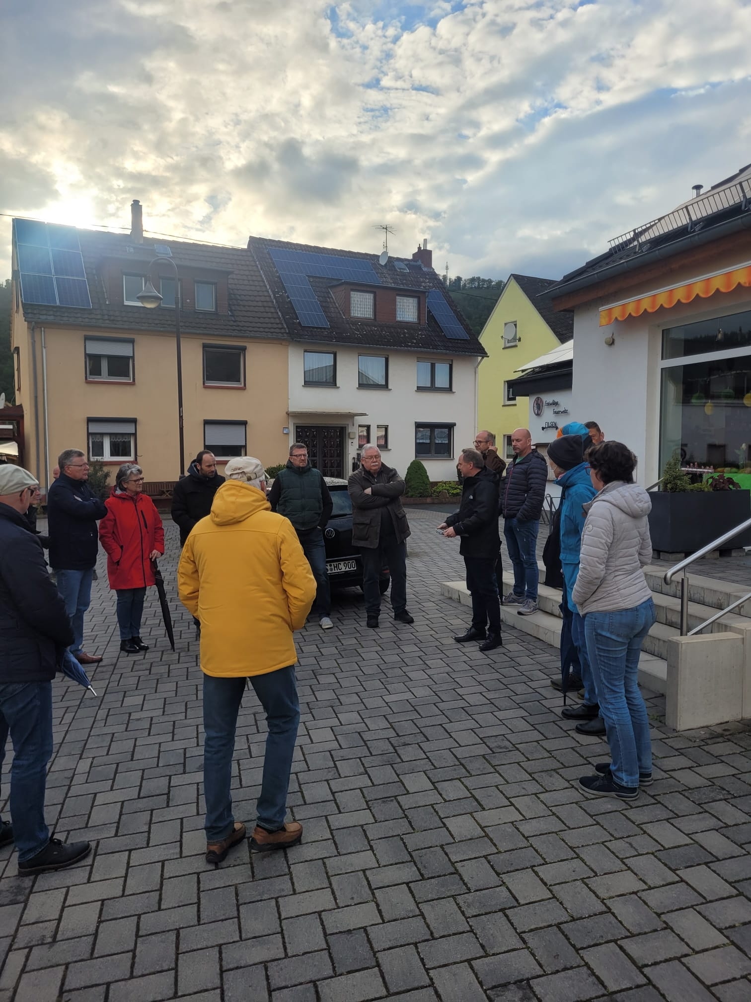 Bürgermeister Mike Weiland und circa 20 weitere Personen stehen bei bewölktem Wetter im Kreis auf dem Dorfplatz in Filsen. 