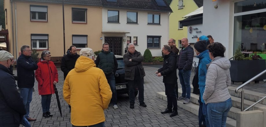 Bürgertreff Filsen Bürgermeister Mike Weiland und circa 20 weitere Personen stehen bei bewölktem Wetter im Kreis auf dem Dorfplatz in Filsen.