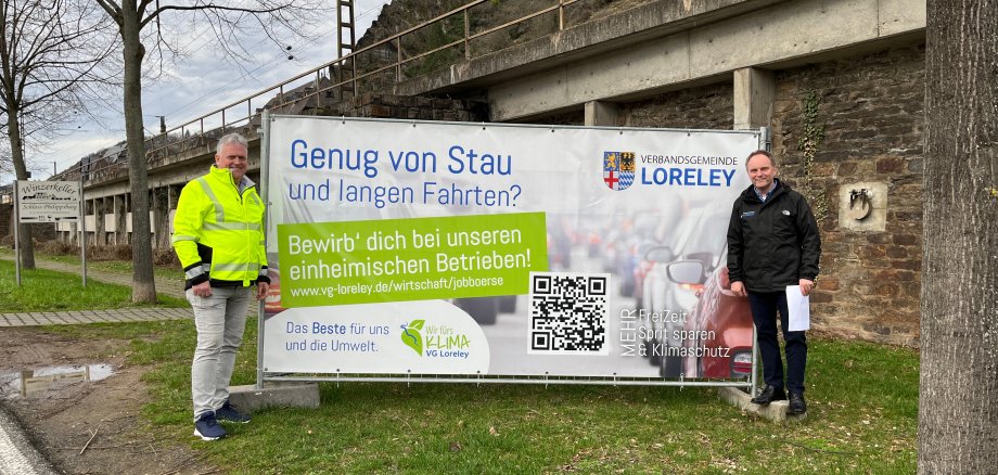 Plakat Jobbörse Herbert Fuss (Abteilungsleiter Verkehr, ADAC Mittelrhein e. V.) und Bürgermeister Mike Weiland stehen vor einem Werbeplakat für die VG-JOBBÖRSE