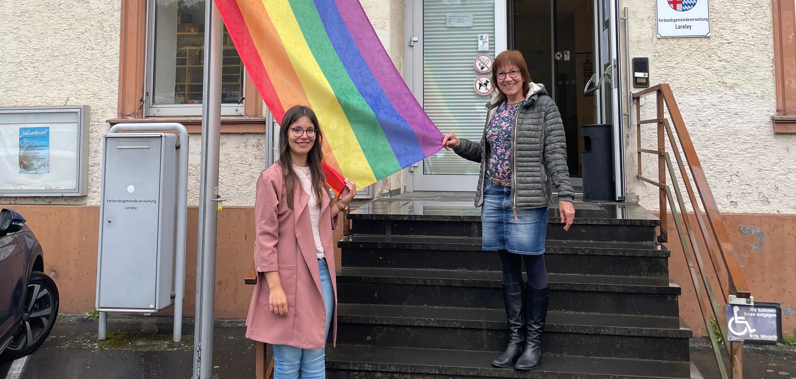 Gleichstellungsbeauftragte Jasmin Fluck und Regionalkoordinatorin nördliches RLP, Frau Laschet-Einig, stehen auf der Treppe vor dem Verwaltungsgebäude in St. Goarshausen und hissen eine Regenbogenflagge.