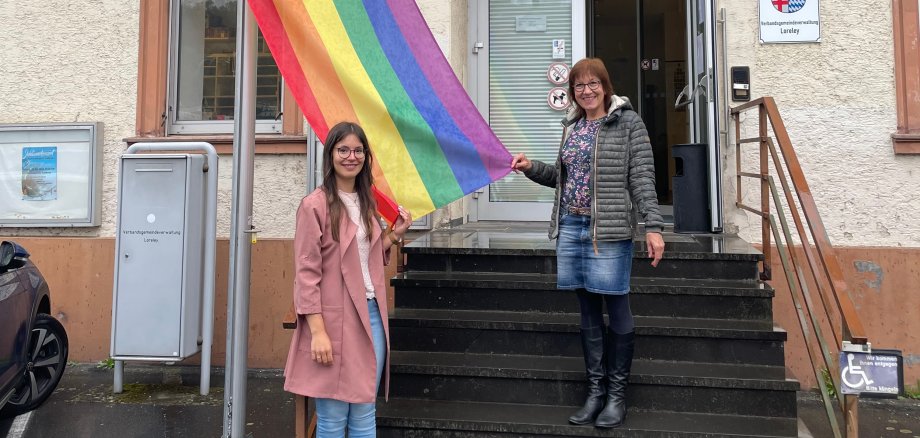 Hissen der Flagge zum Idahobit 2024 Gleichstellungsbeauftragte Jasmin Fluck und Regionalkoordinatorin nördliches RLP, Frau Laschet-Einig, stehen auf der Treppe vor dem Verwaltungsgebäude in St. Goarshausen und hissen eine Regenbogenflagge.