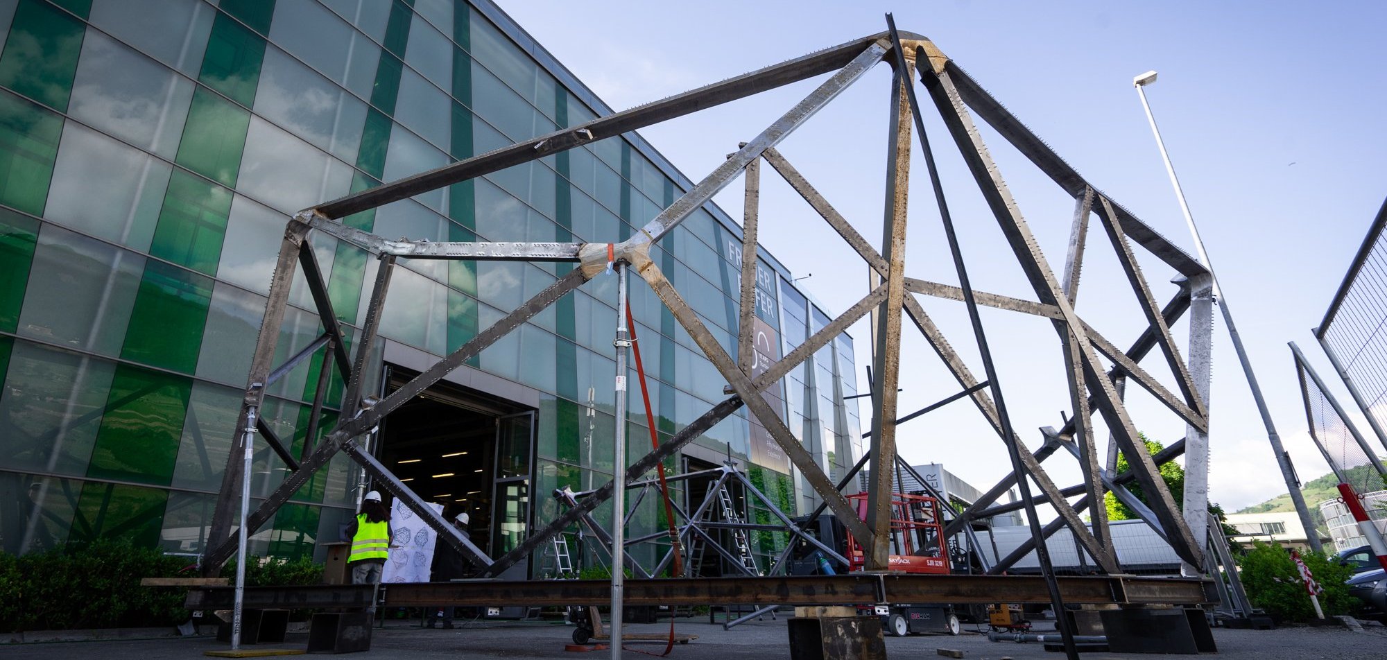Zu sehen ist eine Stahlkonstruktion, die in ihrer Form an einen Felsen erinnert. Diese steht draußen vor einem großen Gebäude.