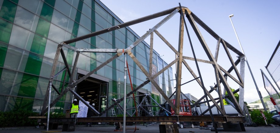Zu sehen ist eine Stahlkonstruktion, die in ihrer Form an einen Felsen erinnert. Diese steht draußen vor einem großen Gebäude.