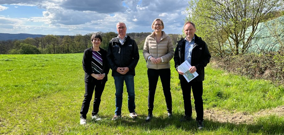 PV VG Loreley von links nach rechts stehen Referatsleiterin Dr. Stefanie Hahn, Bauamtsleiter Gerd Schuster, Staatssekretärin Simone Schneider und Bürgermeister Mike Weiland an einer potenziellen Fläche zur Errichtung einer Photovoltaik-Freiflächenanlage. Das Wetter ist sonnig und im Hintergrund sind Bäume und Berge zu sehen.