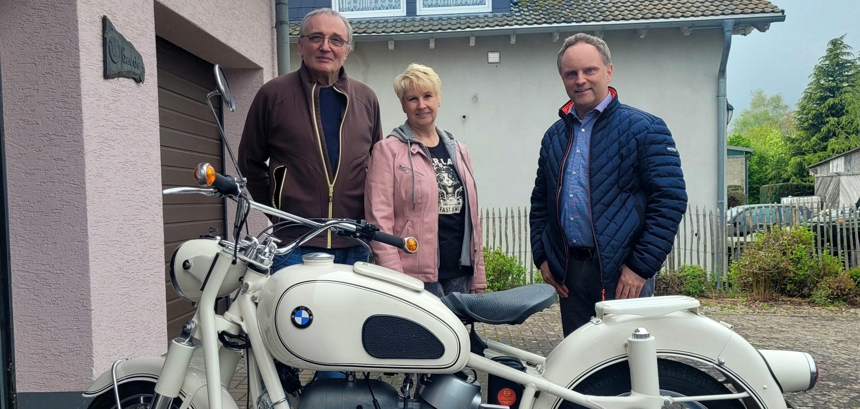 Museumsinhaber Reinhard Gaede, Gisela Adam und Bürgermeister Mike Weiland stehen hinter einem weißen BMW-Oldtimermotorrad. Im Hintergrund ist ein Haus mit Garten zu sehen.