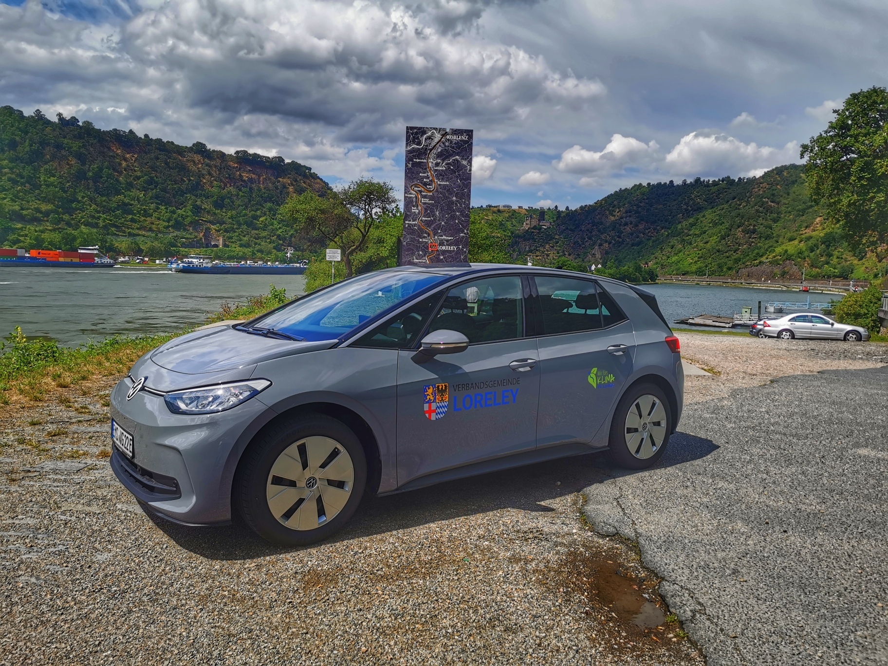 Ein dunkelgrauer VW ID 3 (E-Auto) mit dem VG-Klima-Logo steht bei sonnigem Wetter am Hafen in St. Goarshausen.